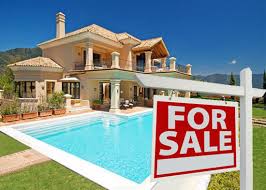 A luxurious beige villa in Spain with a large swimming pool and manicured lawn, under a clear blue sky. A red and white "FOR SALE" sign is prominently displayed in the foreground.